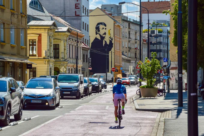 Cyclists in casual spring attire ride along dedicated and protected cycling infrastructure lined with buildings, trees, seating, light fixtures, and grass tramways in Bratislava.
