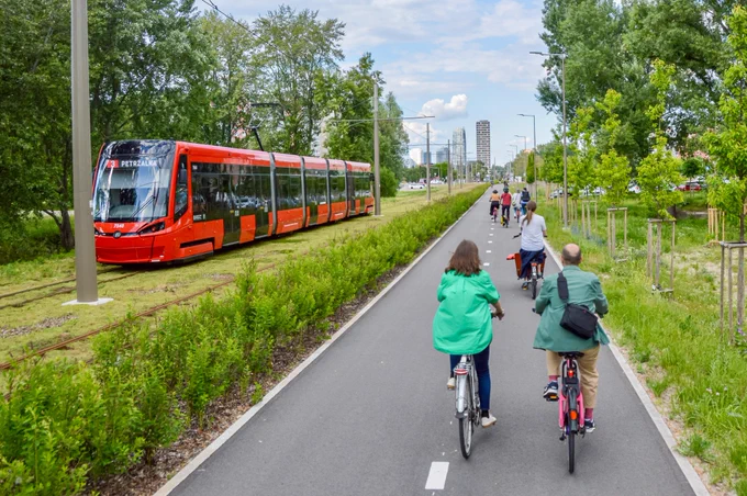 Cyclists in casual spring attire ride along dedicated and protected cycling infrastructure lined with buildings, trees, seating, light fixtures, and grass tramways in Bratislava.