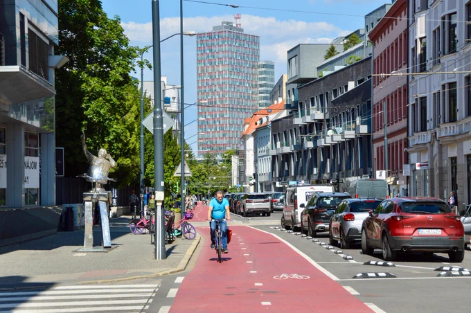 Cyclists in casual spring attire ride along dedicated and protected cycling infrastructure lined with buildings, trees, seating, light fixtures, and grass tramways in Bratislava.