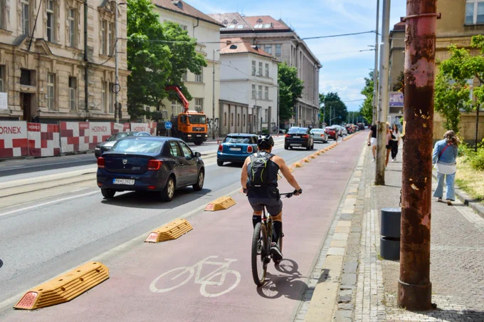 Cyclists in casual spring attire ride along dedicated and protected cycling infrastructure lined with buildings, trees, seating, light fixtures, and grass tramways in Bratislava.