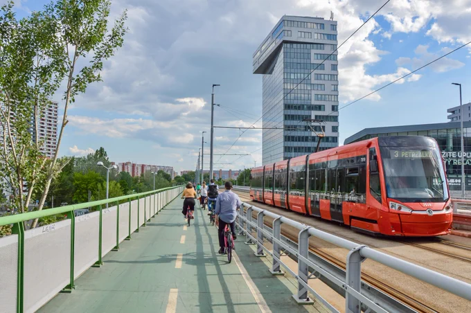 Cyclists in casual spring attire ride along dedicated and protected cycling infrastructure lined with buildings, trees, seating, light fixtures, and grass tramways in Bratislava.
