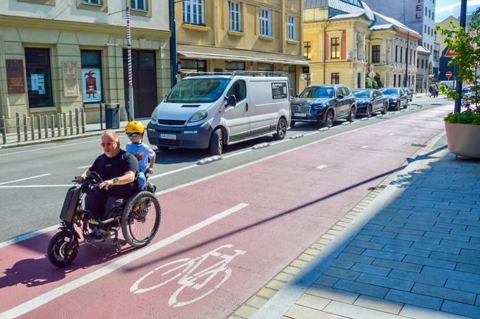 Cyclists in casual spring attire ride along dedicated and protected cycling infrastructure lined with buildings, trees, seating, light fixtures, and grass tramways in Bratislava.