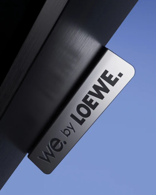Metal sign reading 'We. by Loewe' mounted on a black TV frame against a clear blue sky.