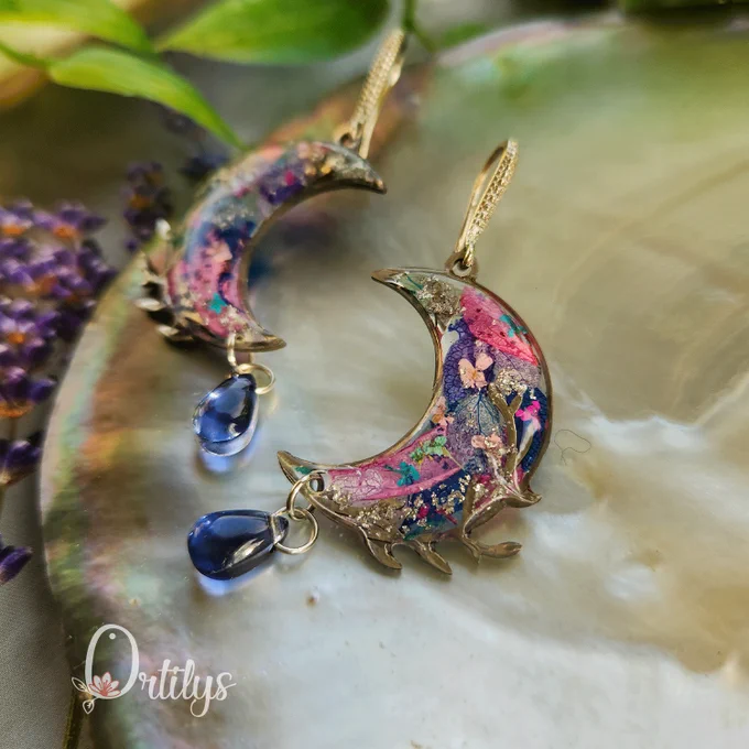 boucles d'oreilles argentées en forme de lune, dans les coloris bleus, violets et roses. une perle bleue pend du croissant de lune