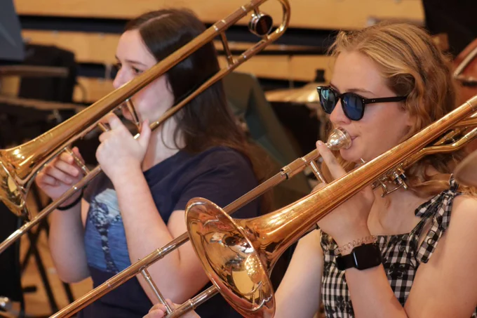 Two young musicians playing trombones in a rehearsal room filled with musical instruments.
