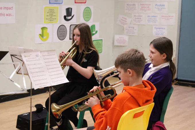 Three children seated in a classroom playing brass instruments with sheet music on stands in front of them.