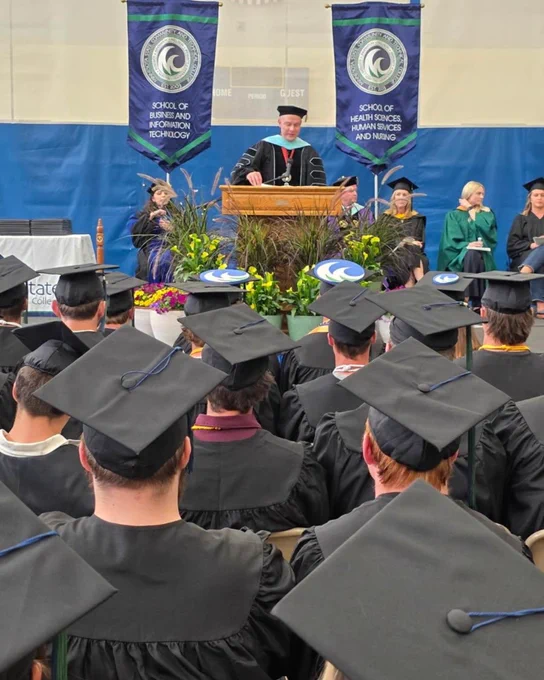 A speaker stands at a podium on a stage, addressing a graduation ceremony. Graduates in caps and gowns are seated in the audience. Two banners with the school's name are displayed behind the speaker.