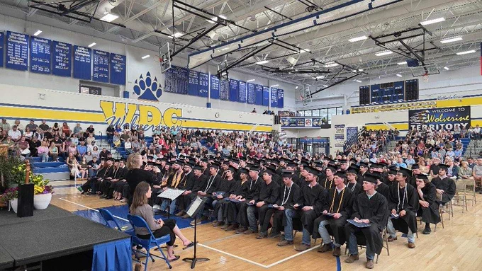 A gymnasium filled with people attending a graduation ceremony. Graduates in black caps and gowns are seated in rows. A podium and speakers are positioned at the front. The wall displays "WDC Wolverines" and banners. An audience watches from bleachers.