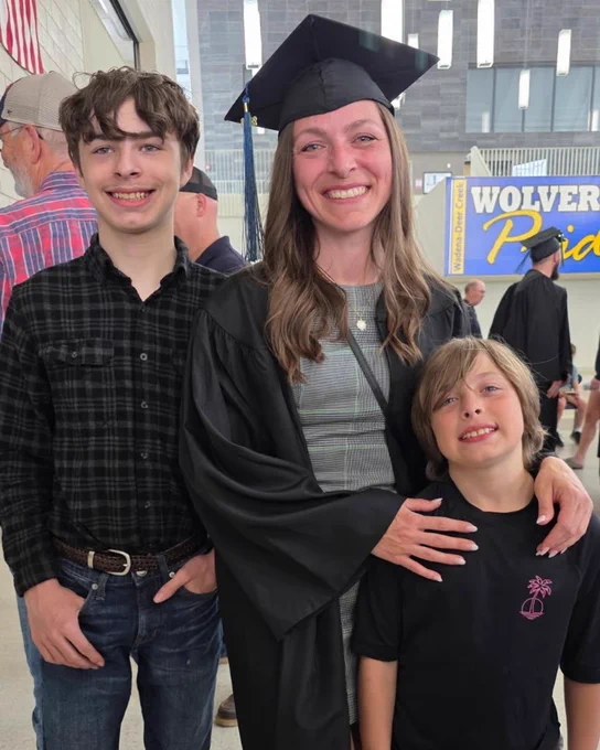 A person in a graduation cap and gown smiles while standing with two children. One child stands on each side. The backdrop includes a sign with the word "Wolverines".