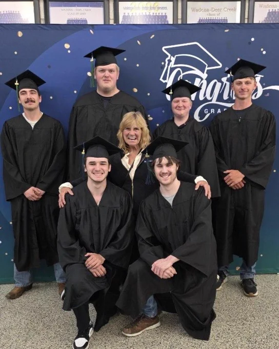 A group of people wearing graduation caps and gowns pose together in front of a backdrop. They are smiling, and one person in casual attire stands in the middle. The backdrop features a blue background with gold dots.