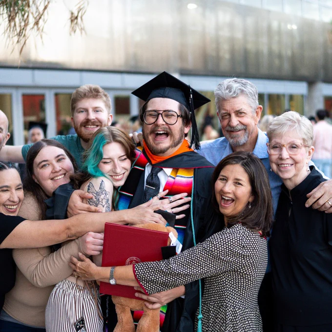 A group celebrates a graduation outside The Pit. The graduate, wearing a cap and gown with colorful stoles, holds a diploma and teddy bear, joyfully surrounded by smiling people in casual attire.