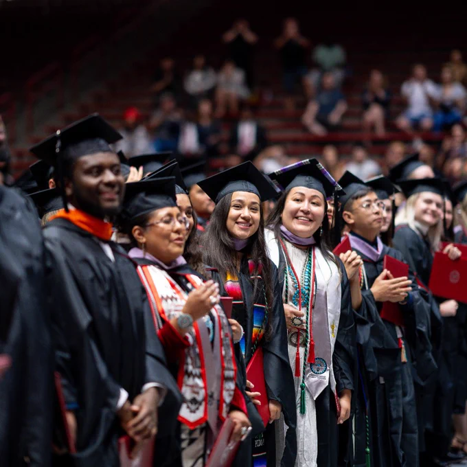 A group of graduates stands in a line wearing caps and gowns. Some hold diplomas, and several wear colorful stoles and cords. The setting is a large auditorium filled with seated spectators.