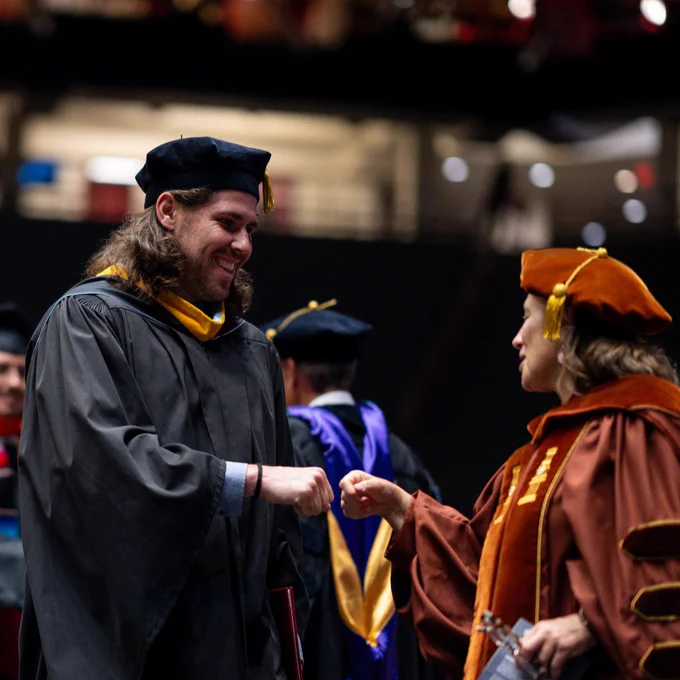 A graduate in academic regalia fist-bumps a faculty member at a commencement ceremony.