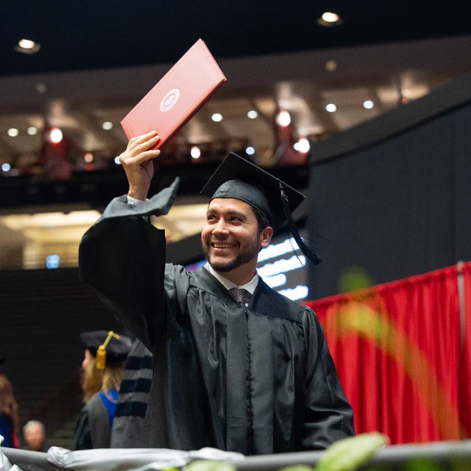 A person in a black graduation gown and cap is smiling and holding up a red diploma cover at a graduation ceremony. The background shows red curtains, an auditorium with people sitting, and decorative plants in the foreground.