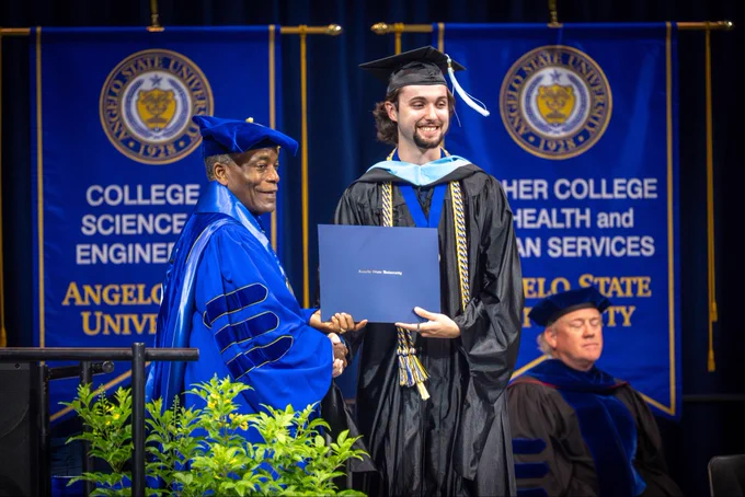 Two individuals pose for a photo at a commencement ceremony. They hold a diploma between them.