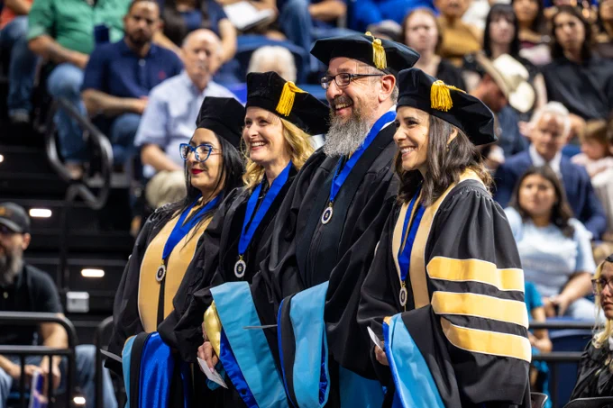 A group of four people in graduation gowns and doctoral tams stand smiling. 