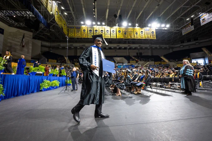 A graduate in a black cap and gown, proudly walks with a diploma folder.