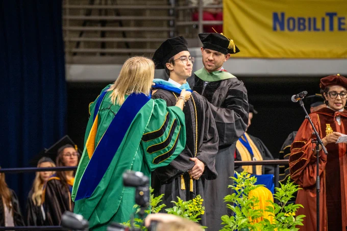 A graduate in a cap and gown is receiving a masters hood by a faculty member.