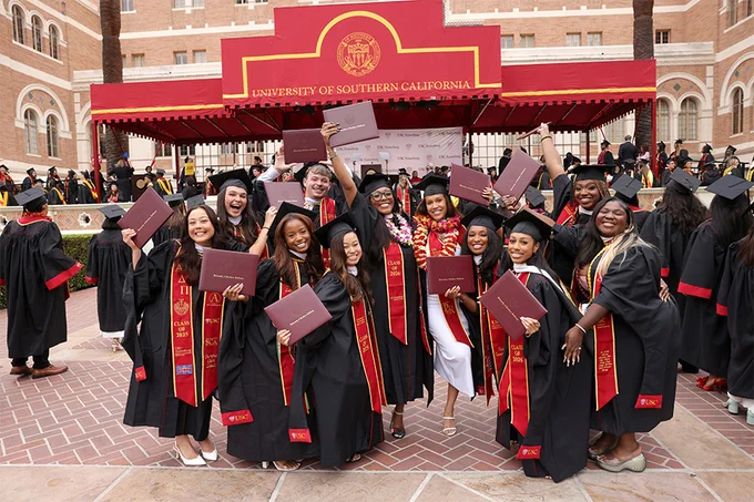 A group of graduates pose together in front of the USC Annenberg commencement stage, holding diplomas and smiling after the ceremony.