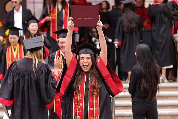 A graduate in cap and gown raises a diploma overhead and smiles in celebration after commencement.