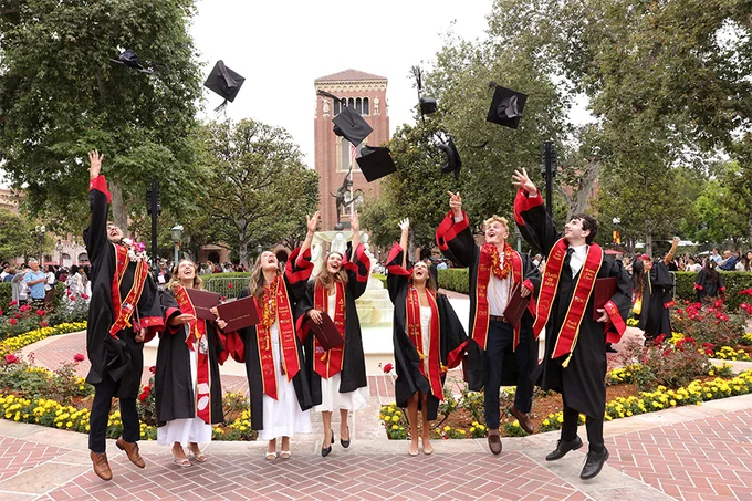 Graduates in black caps and gowns toss their mortarboards into the air in front of a campus fountain and Bovard Auditorium during USC Annenberg’s commencement ceremony.
