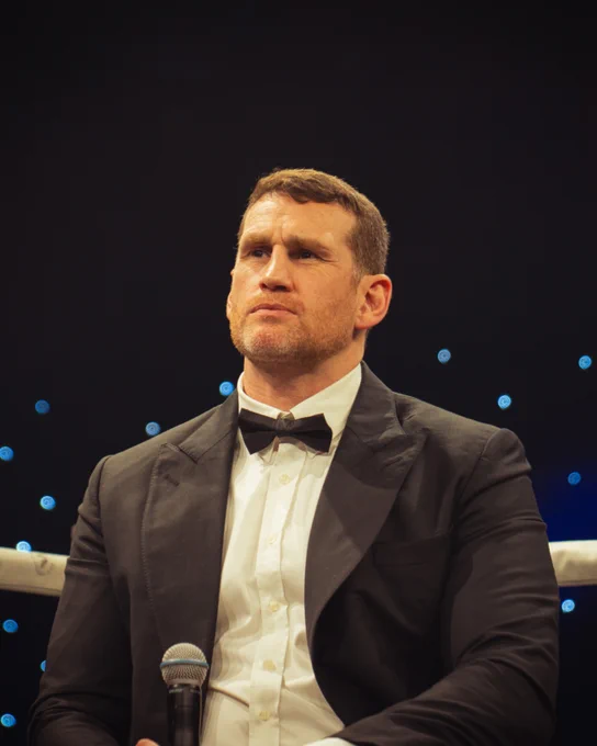 A man dressed in a tuxedo sits with a microphone, appearing contemplative. The background is dark with small blue lights, adding a formal ambiance.