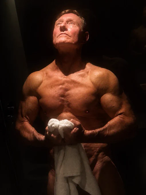 Portrait artistique d’un homme de 63 ans à la musculature athlétique sous une lumière chaude et dramatique après l’effort, tenant une serviette dans une ambiance sombre. 