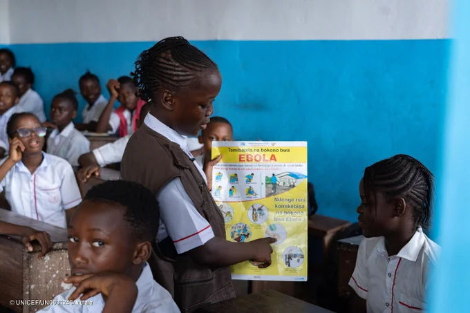 Une Enfant Reporter discute avec une élève dans une salle de classe, en montrant du doigt une affiche marquée "Ebola".