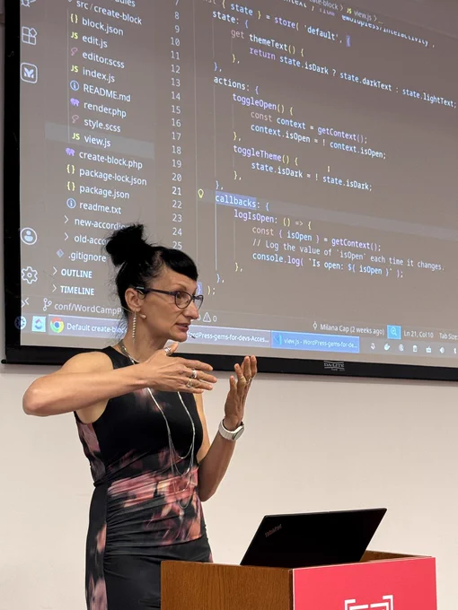 A woman with glasses and dark hair tied up speaks in front of a projection screen displaying code in a VS Code interface during a presentation.