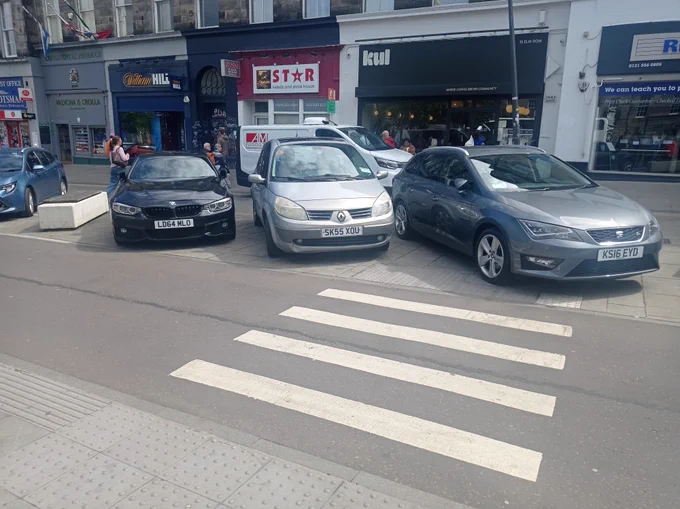 zebra on cycle lane to the shops. Cars parked on pavement and tactiles