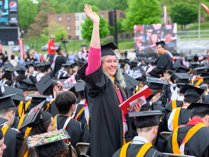 A graduate wearing a cap and gown stands and waves during an outdoor graduation ceremony. The person holds a red folder and smiles among seated graduates. In the background, there's a score display with a Pepsi logo and green trees.