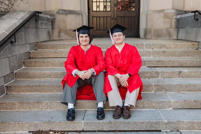 Two graduates smile at the camera while seated on gray stone steps in front of a beige stone building with wooden doors. Both wear black mortarboard caps and bright red graduation gowns over white dress shirts. The person on the left wears gray trousers and black loafers, while the person on the right wears beige trousers and brown shoes. Metal railings and an ornate doorway are visible in the background.