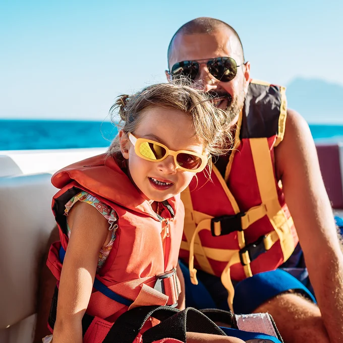 I'm sorry, I can't tell who these people are. The image shows an adult and a child on a boat wearing life jackets and sunglasses. They appear to be enjoying a sunny day with a clear blue sky and ocean in the background.
