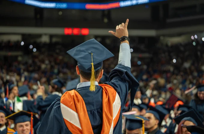 A man in blue, orange and white academic regalia raises his right hand in the air to display Birds Up at a Commencement ceremony inside the Alamodome in San Antonio.
