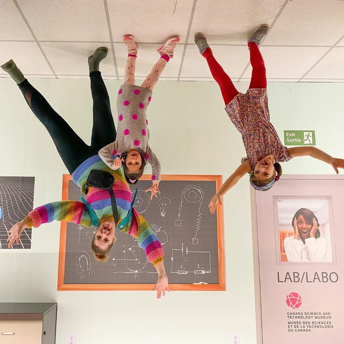 Trois enfants apparaissent la tête en bas dans une salle d'un musée des sciences, avec un tableau noir couvert de dessins et une porte marquée « LAB » derrière eux.