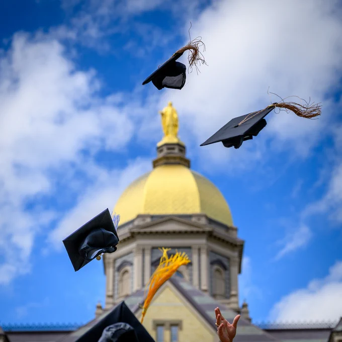 Graduation caps are tossed in the air in front of the University of Notre Dame's iconic Golden Dome, set against a blue sky with clouds.