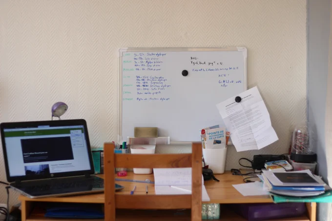 Sur un bureau, un ordinateur portable, des cours, et un petit tableau blanc avec le programme de la semaine, et accroché à l'aide d'un aimant, une attestation de sortie dérogatoire utilisée durant le premier confinement dû au COVID-19.