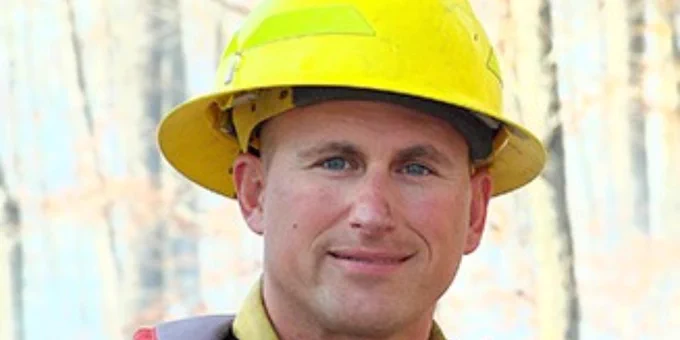 Construction worker wearing a bright yellow hard hat and safety glasses at an outdoor site with blurred trees in the background.