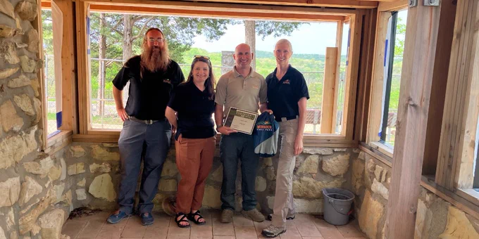 Four people standing inside a stone-walled room with large windows overlooking a green landscape, one holding a certificate and a gift bag.