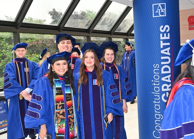 A group of people in graduation attire stand smiling in a hallway. They are wearing blue robes with hoods, and one of them has a colorful stole. A banner reading "Congratulations Graduates" with the American University logo is visible.