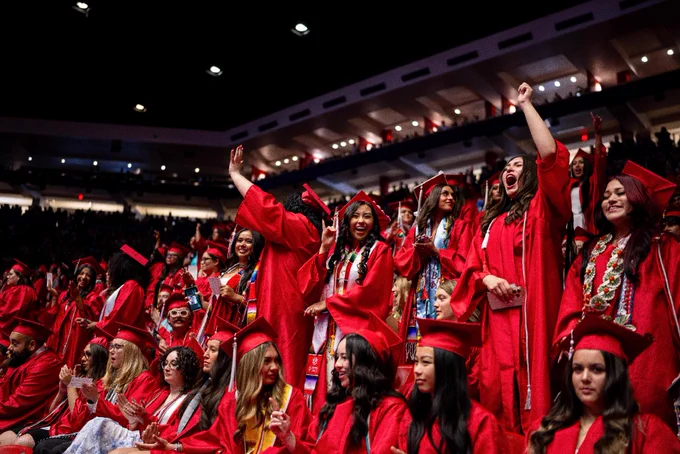 A large group of graduates in red caps and gowns celebrate in an indoor arena. The crowd is lively, with some individuals standing and raising their hands in excitement. The arena is filled with people, and the atmosphere is festive.