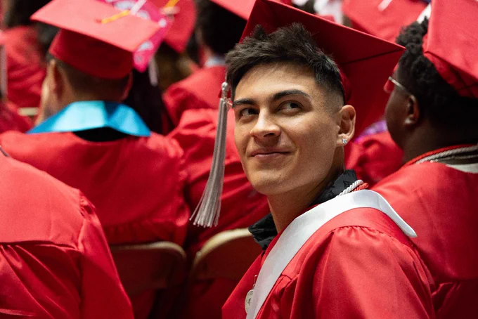 A person in a red graduation gown and cap looks over their shoulder, surrounded by other graduates in similar attire. Some caps are decorated with colorful designs. 