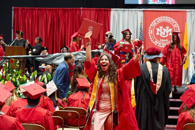 A person wearing a graduation cap and gown excitedly holds up a diploma in a crowded graduation ceremony. The backdrop features the UNM emblem. Other participants in red gowns and faculty members are visible.