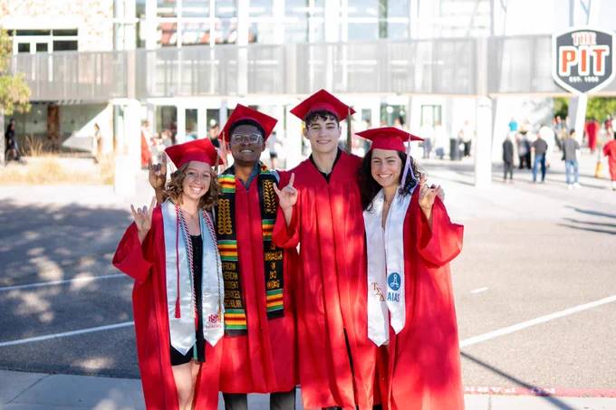 A group of four people in red graduation gowns and caps stand together, smiling in front of The Pit. Two wear stoles decorated with colorful designs.
