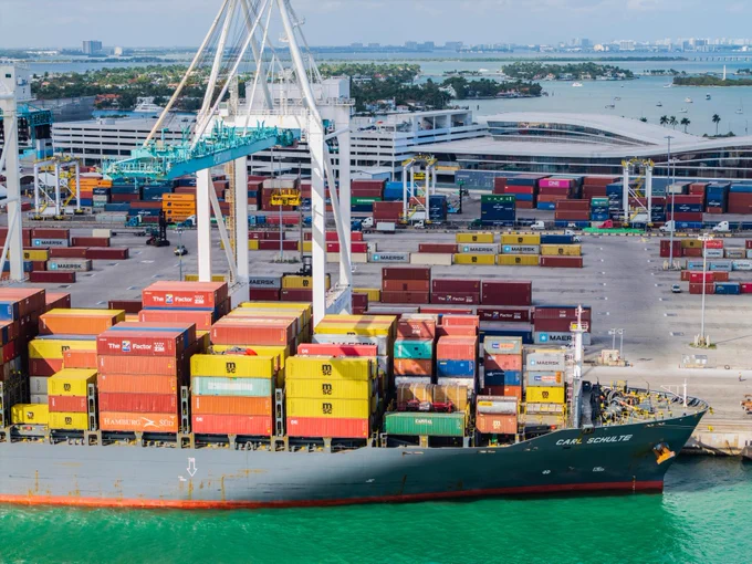 Cargo ship \"CARL SCHULTE\"@1 docked at a port, loaded with colorful shipping containers. Cranes move containers in the background near buildings and a bay with boats. Text includes \"MAERSK\"@2, \"The Factor ZIM\"@3, \"HAMBURG S\u00DCD\"@4, \"CAPITAL\"@5.