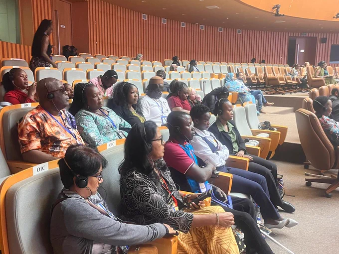 A diverse group of people wearing headsets sit attentively in a spacious auditorium with wooden walls, conveying focus and engagement during the review of state report of Sarhawi Democratic Republic
