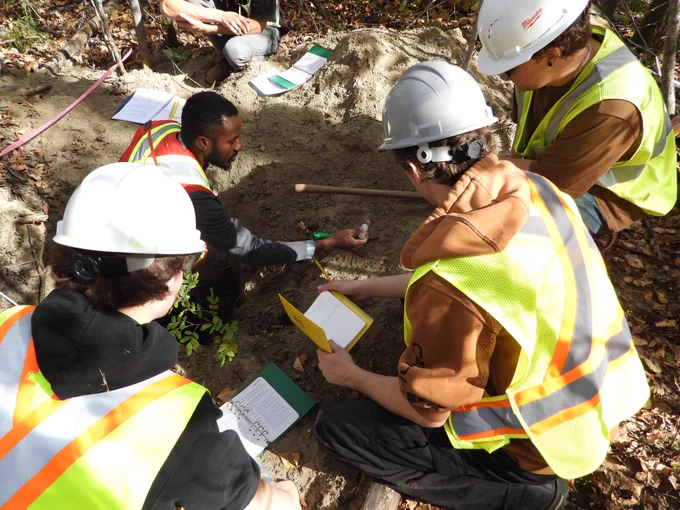 Dr. Emmanuel Badewa teaching students how to characterize a forest soil. Credit: David Raitt    ALT Four men, wearing reflective vests and hard hats, in a soil pit observing soil