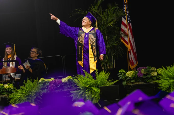 A graduate smiles and points to the crowd as they walk across the stage during a commencement ceremony.