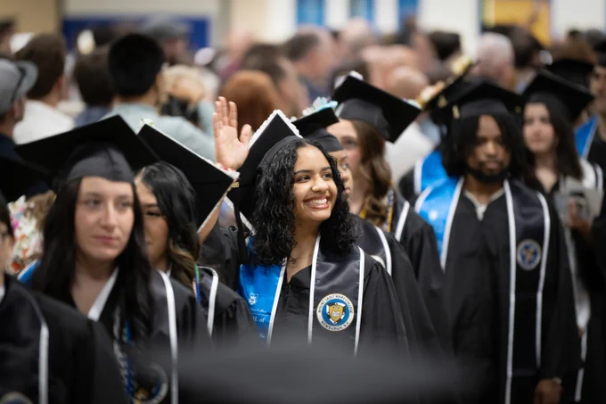 Graduates wearing their graduation regalia. One graduate is waving.
