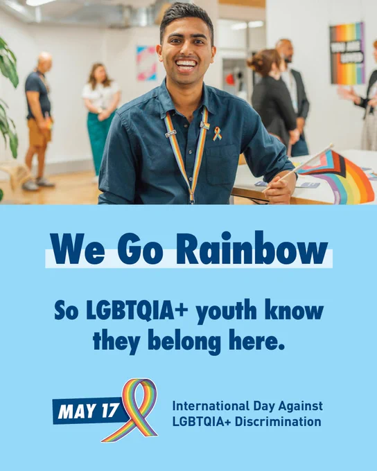 A man is smiling and wearing a rainbow lanyard and ribbon while holding a rainbow flag. He is in an office setting with colleagues. Below text says 'We Go Rainbow. So LGBTQIA  youth know they belong here.' Below it says 'International Day Against LGBTQIA  Discrimination. May 17.' 